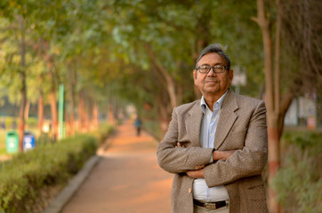Fototapeta premium Portrait of a happy senior Indian man wearing a tweet suit/coat and hands crossed in the outside setting standing and thinking in a park in winters in New Delhi, India