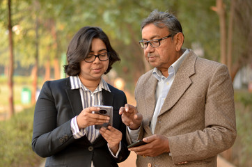 Young Indian woman manager/entrepreneur in western formals or suit with old Indian man. Both working on their own mobile promoting digital literacy for elderly in a park in New Delhi, India
