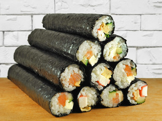 Uncut simple homemade sushi rolls stuffed with crab sticks, cucumber, salted salmon, soft cheese and omelet on wooden surface on background of white brick wall.