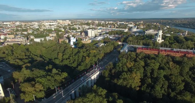 An aerial view of Kaluga urbanscape with multi storey modern and historical buildings and a bridge in the middle of the view. Green tree crowns are filling spaces between buildings. Blue sky and river