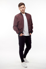 Young european man in white sweater and black pants, red bomber jacket posing on white background....