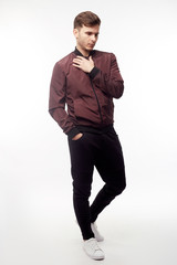 Young european man in white sweater and black pants, red bomber jacket posing on white background....