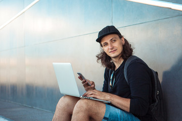 A young man, sitting on a skateboard, is trying to synchronize a laptop with a phone. Look into the...