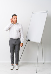 Young latin coaching man with a whiteboard isolated trying to listening a gossip.