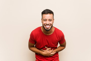 Young south-asian man laughs happily and has fun keeping hands on stomach.