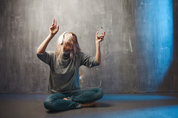 Positive young blond woman in a grey oversized sweater dancing to the music in headphones