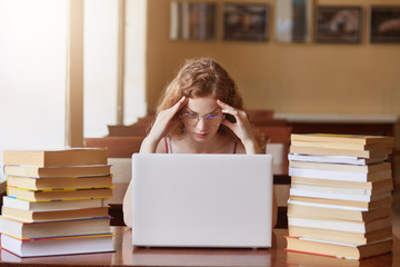 Young tired student being in university library, looking at her lap to pscreen, keeping hands on...