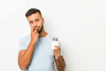 Young south-asian man holding an after shave cream relaxed thinking about something looking at a copy space.