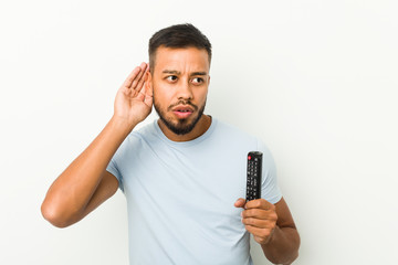 Young south-asian man holding a tv controller trying to listening a gossip.