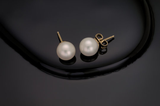A close up of a pair of white pearl earring studs with yellow gold post and nut on a black reflective curved melted step background.