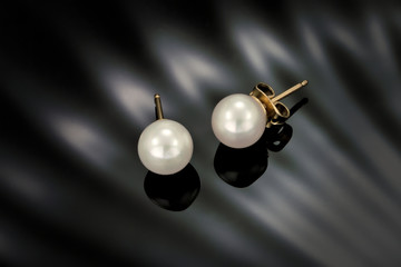 A close up of a pair of white pearl earring studs with yellow gold post on a black fluted refective background.