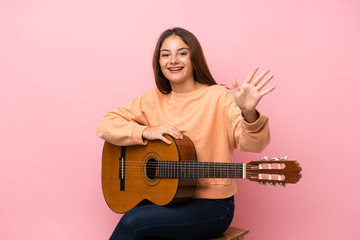 Young brunette girl with guitar over isolated pink background saluting with hand with happy expression