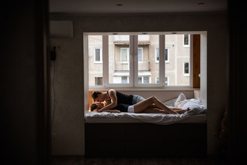 Panoramic view of a modern apartment with a window near which a couple in love hugs on the bed. Lovers with tenderness rest on a festive day off at home. The concept of tactility, trust in a partner