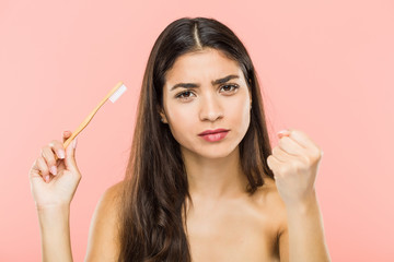 Young indian woman holding a toothbrush showing fist to camera, aggressive facial expression.