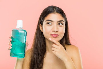 Young indian woman holding a mouthwash bottle looking sideways with doubtful and skeptical expression.