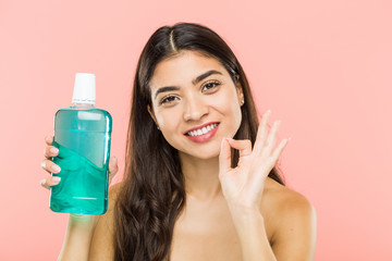 Young indian woman holding a mouthwash bottle cheerful and confident showing ok gesture.