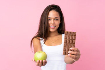 Young Brazilian girl over isolated pink background taking a chocolate tablet in one hand and an...