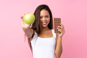Young Brazilian girl over isolated pink background taking a chocolate tablet in one hand and an...
