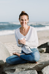 Woman Sitting By The Ocean Holding A Cup And Smiling, Relaxed Yoga Lifestyle Concept