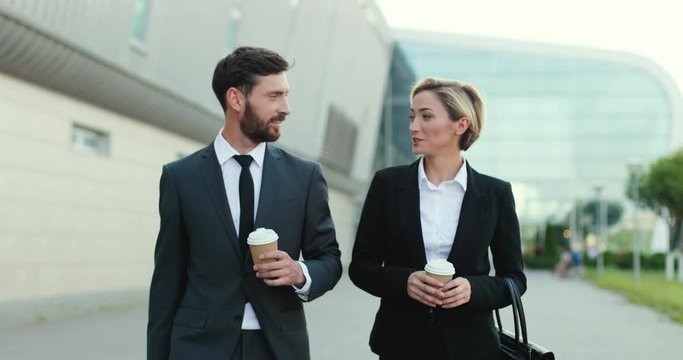 Cheerful attractive and stylish Caucasian business couple walking the street together in the morning, having a nice conversation and carrying cups with coffee to-go in hands while it's hot.