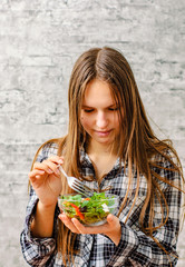 portrait of young teenager brunette girl with long hair eating green vegetables salad on gray wall background