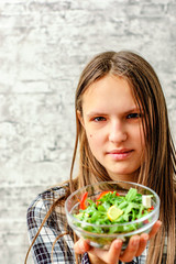 portrait of young teenager brunette girl with long hair eating green vegetables salad on gray wall background