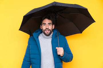 Man holding an umbrella over isolated yellow background with surprise and shocked facial expression