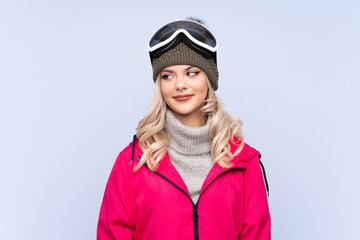 Skier teenager girl with snowboarding glasses over isolated blue background standing and looking to the side