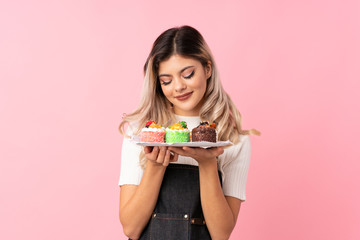 Teenager girl over isolated pink background holding mini cakes enjoying the smell of them