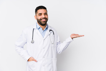 Young arabian doctor man isolated showing a copy space on a palm and holding another hand on waist.