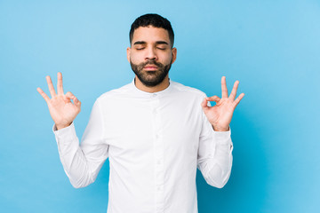 Fototapeta premium Young latin man against a blue background isolated relaxes after hard working day, she is performing yoga.