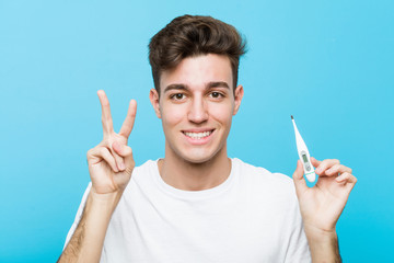 Young caucasian man holding a medical thermometer showing number two with fingers.