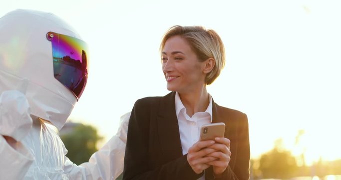 Young Caucasian beautiful woman tapping on the phone while funny astronaut going around her and looking at the screen what she doing. Outside. Close up.