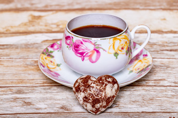 A white porcelain Cup on a saucer with a delicate pattern of pink and yellow rose buds with instant coffee on a wooden background with a heart-shaped chocolate gingerbread.