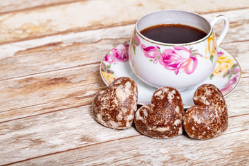 A white porcelain Cup on a saucer with a delicate pattern of pink and yellow rose buds with instant coffee on a wooden background with a heart-shaped chocolate gingerbread.