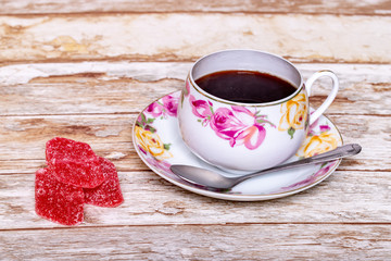 White porcelain Cup with a delicate pattern of pink and yellow rose buds with instant coffee with a spoon and pieces of marmalade on a saucer on a wooden background.