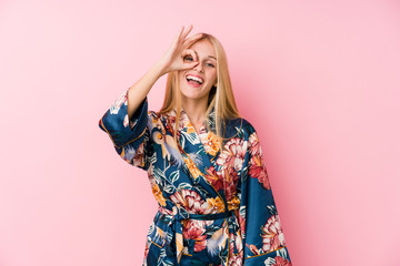 Young blonde woman wearing a kimono pajama excited keeping ok gesture on eye.