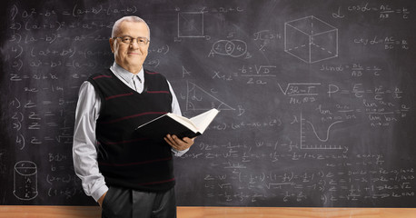 Elderly male teacher holding a book and standing in front of a blackboard with math