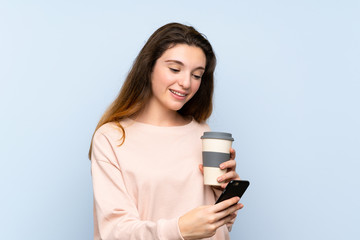 Fototapeta premium Young brunette girl over isolated blue background holding coffee to take away and a mobile