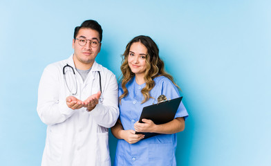 Young doctor couple posing in a blue background isolated holding something with palms, offering to camera.