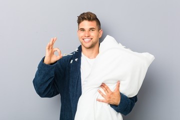 Young man wearing pijama holding a pillow cheerful and confident showing ok gesture.