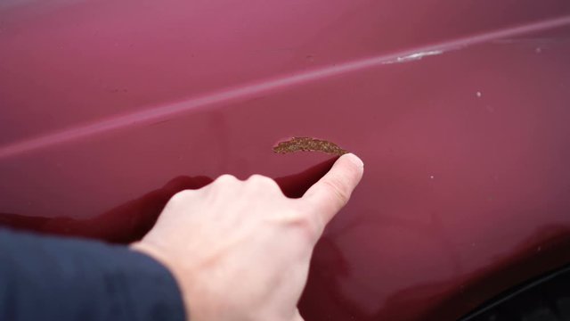 Man shows scratches with rust in a car paint. Finger show scratch on red car