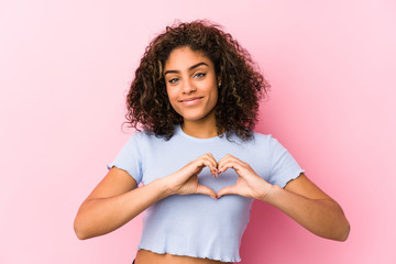 Young african american woman against a pink background smiling and showing a heart shape with hands.