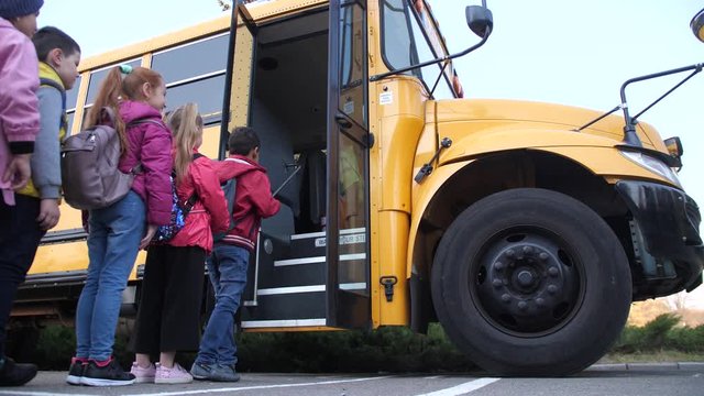Lined up little pupils getting on yellow school bus to go home after lessons. Doors of big school bus opening to let in preadolescent schoolchildren obediently waiting in line to enter inside
