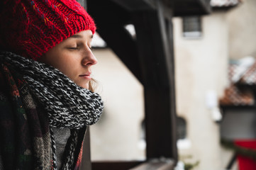 Beautiful young woman with eyes closed, relaxed, thinking and admiring the landscape from a balcony with winter clothes, hat, scarf and coat