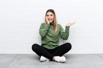 Young caucasian woman sitting on the floor holds copy space on a palm, keep hand over cheek. Amazed and delighted.