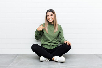 Young caucasian woman sitting on the floor person pointing by hand to a shirt copy space, proud and confident