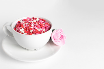 white cup of coffee on a white background filled with heart shaped confetti. pink rose lies on the saucer selective focus. valentine day concept.