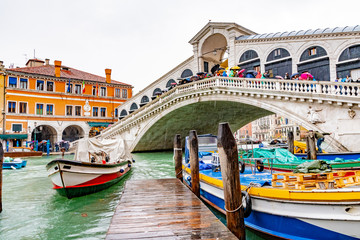 Fototapeta na wymiar Rainy November day/ season in Venice, Italy. People/ tourists walk/ stand with umbrellas on Rialto Bridge on Grand Canal. Colorful boats docked and sailing on water below. Famous landmark attraction.