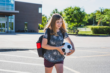 Happy teen girl/student walking to her school in the morning while wearing a backpack and holding a soccer ball.